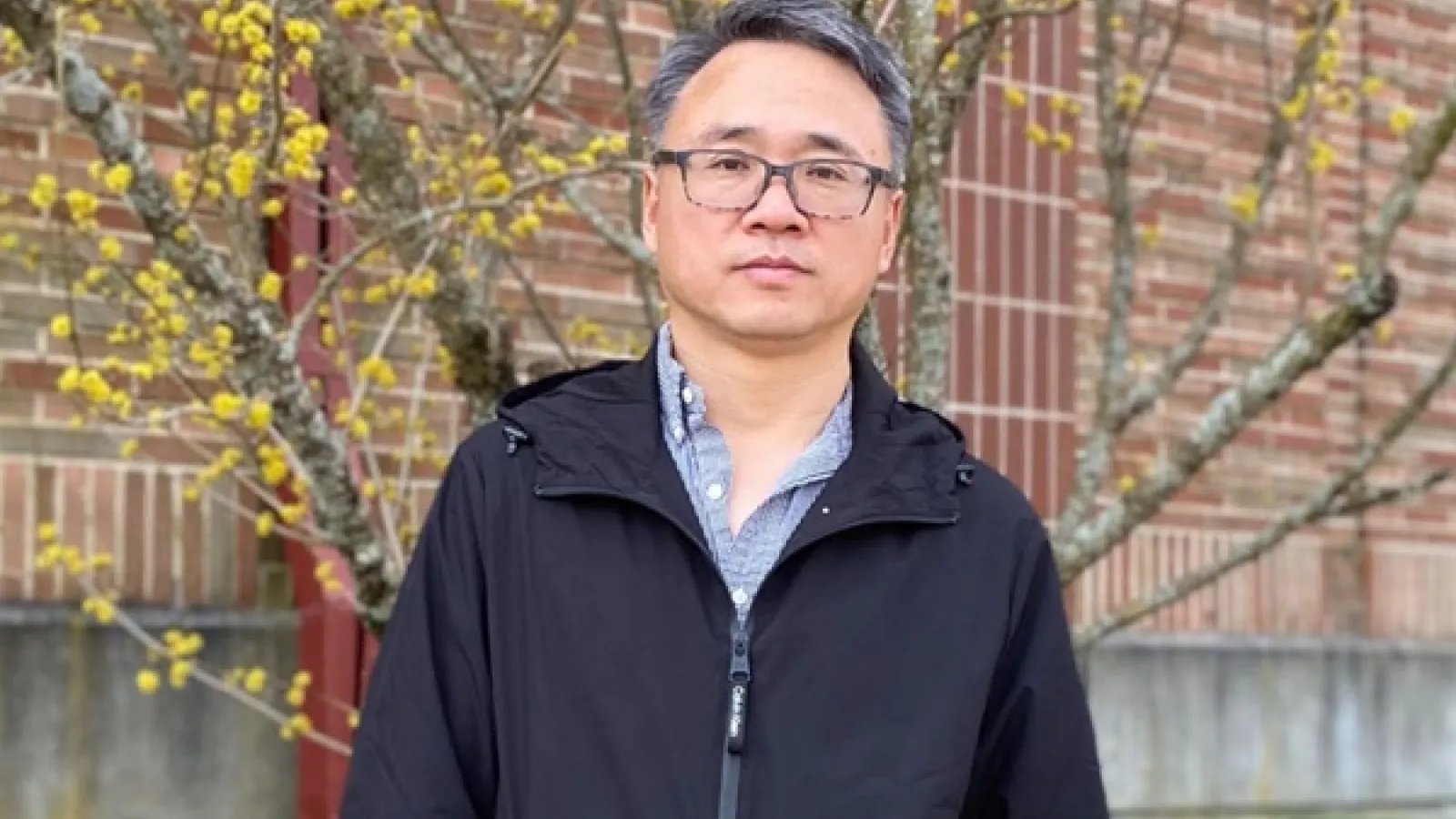  A man wearing glasses and a black jacket stands outdoors in front of a brick wall and a tree with yellow buds. 