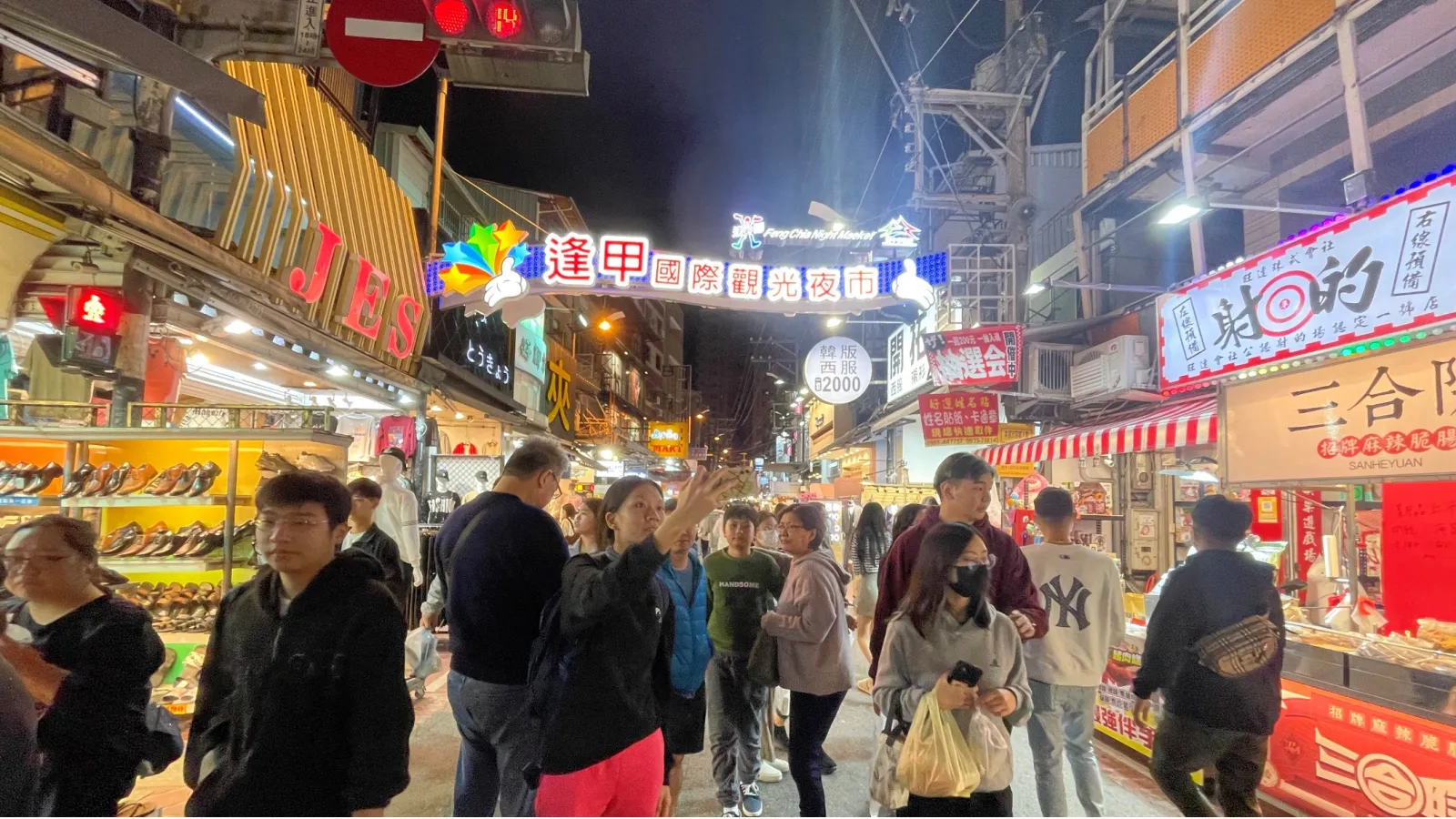  Crowds walk through a brightly lit night market lined with shops and food stalls, with colorful signs in Chinese visible above. 