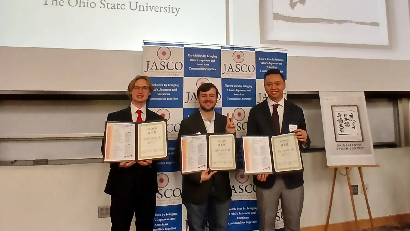  Three men holding certificates stand in front of a display for the 27th Ohio Japanese Speech Contest at The Ohio State University. 