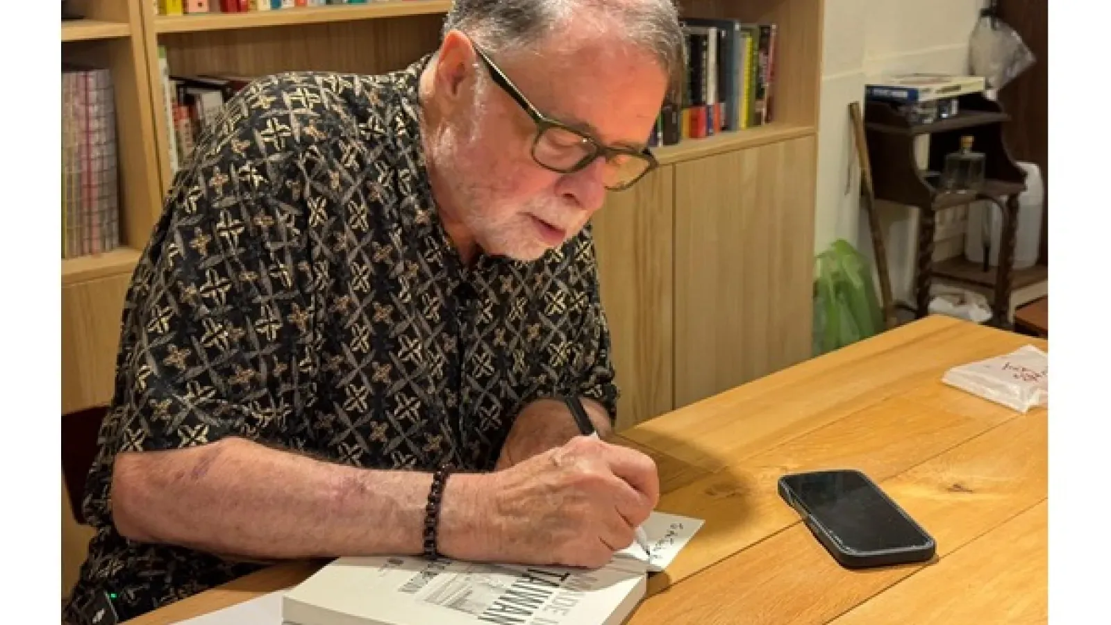 Older man writing beside an open book at a wooden table, with a smartphone nearby and bookshelves in the background.
