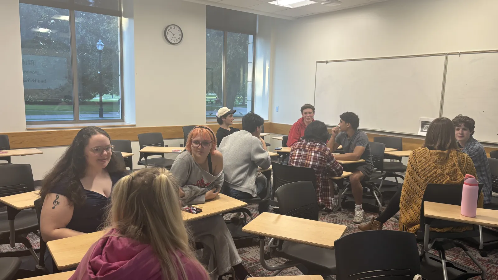 Students sit and converse in a classroom with desks, large windows, a ceiling-mounted projector, a wall display, and an analog clock.