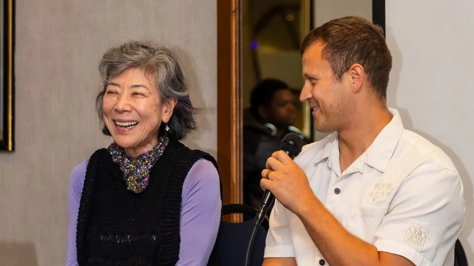 older woman and young man sitting in chairs next to each other, on a stage and addressing a small crowd