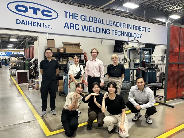  Eight people pose for a group photo inside a factory, standing and kneeling in front of a sign reading "The Global Leader in Robotic Arc Welding Technology. 