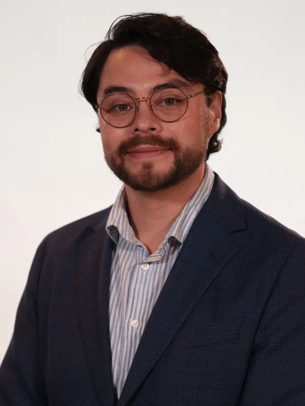 A man with glasses and a beard is wearing a dark blazer over a striped shirt, standing against a plain, light-colored background.