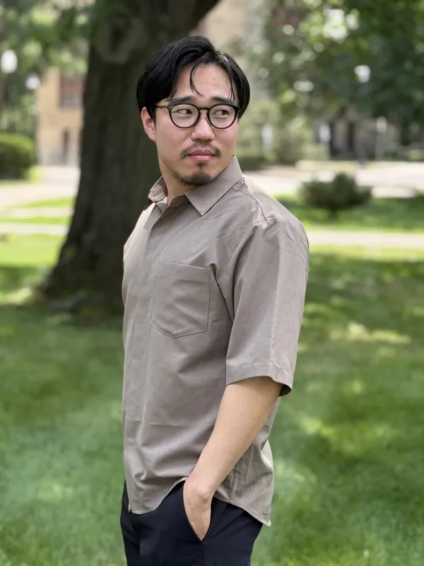 A man wearing glasses and a short-sleeved beige shirt stands outdoors with one hand in his pocket.