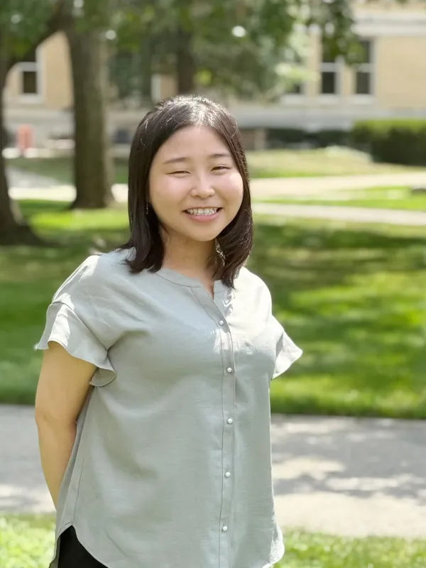 A woman with straight dark hair, wearing a light grey short-sleeve blouse, stands outdoors on a sunny day with grass, trees, and a building in the background.