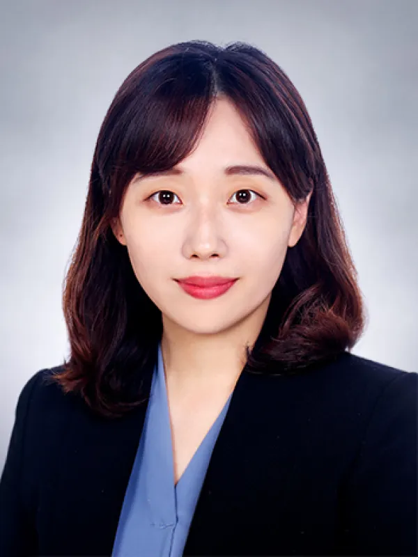 A woman with shoulder-length brown hair, wearing a black blazer over a blue top, poses in front of a plain light gray background.
