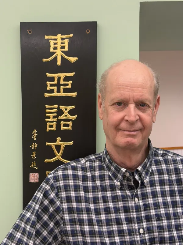 A man in a checkered shirt stands smiling beside a sign with gold East Asian script on a black background. The setting is casual and academic.