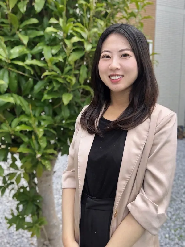 A woman in a black shirt and blazer stands confidently in front of a large green tree. 
