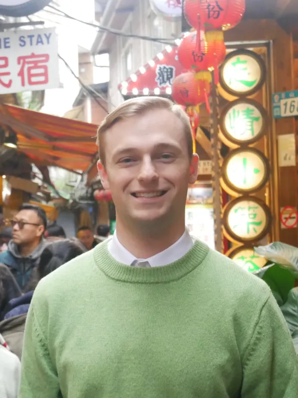A young man in a green sweater smiles in a busy street market with red lanterns and signs in English and Chinese.