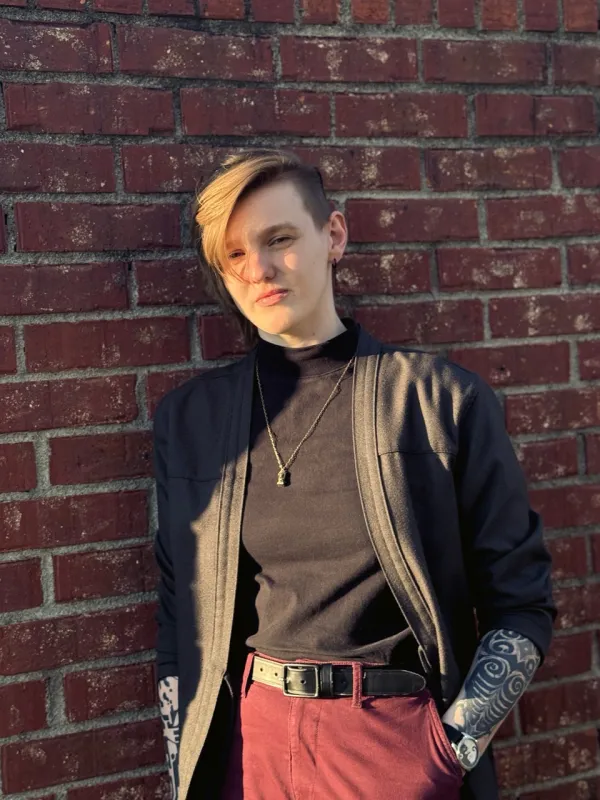 A person in a black jacket stands casually against a brick wall